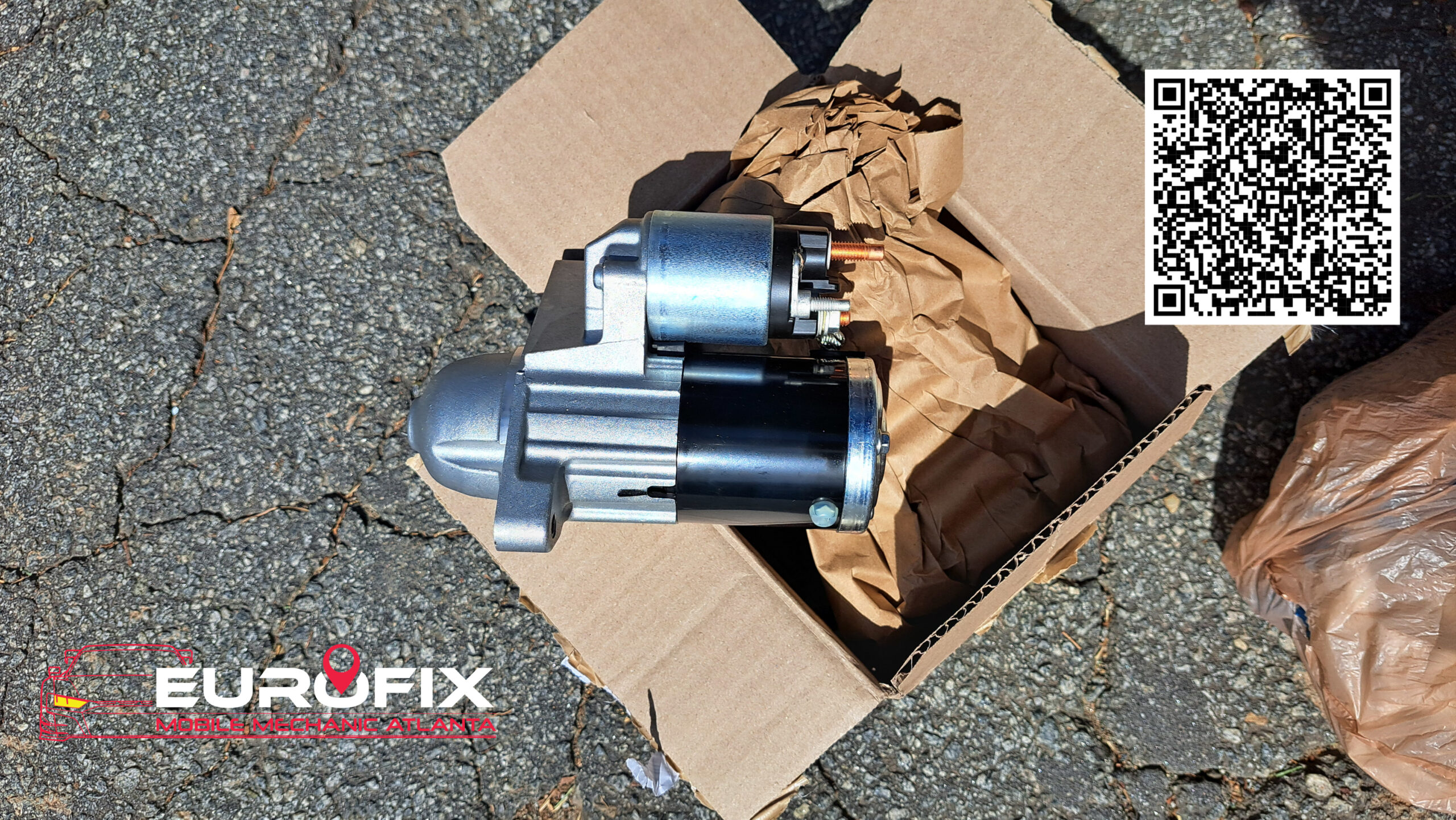 New automotive starter motor sitting in an open cardboard box on pavement, ready for installation.